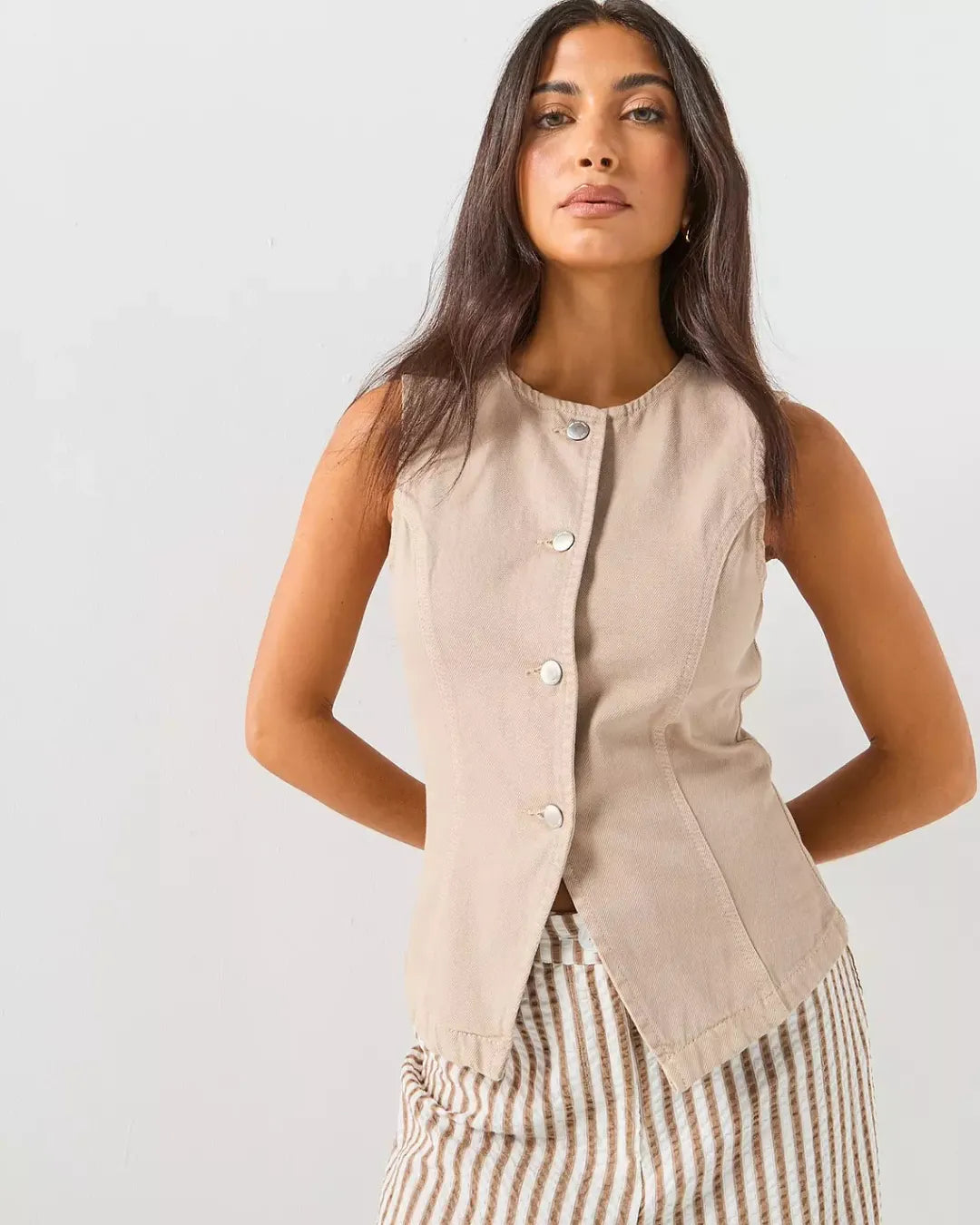 Front view of a woman wearing ONLY beige brown sleeveless waistcoat top with metallic buttons and striped trousers against a plain background
